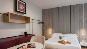 A modern hotel room with a bed, bedside table, chair, and a desk with a computer.