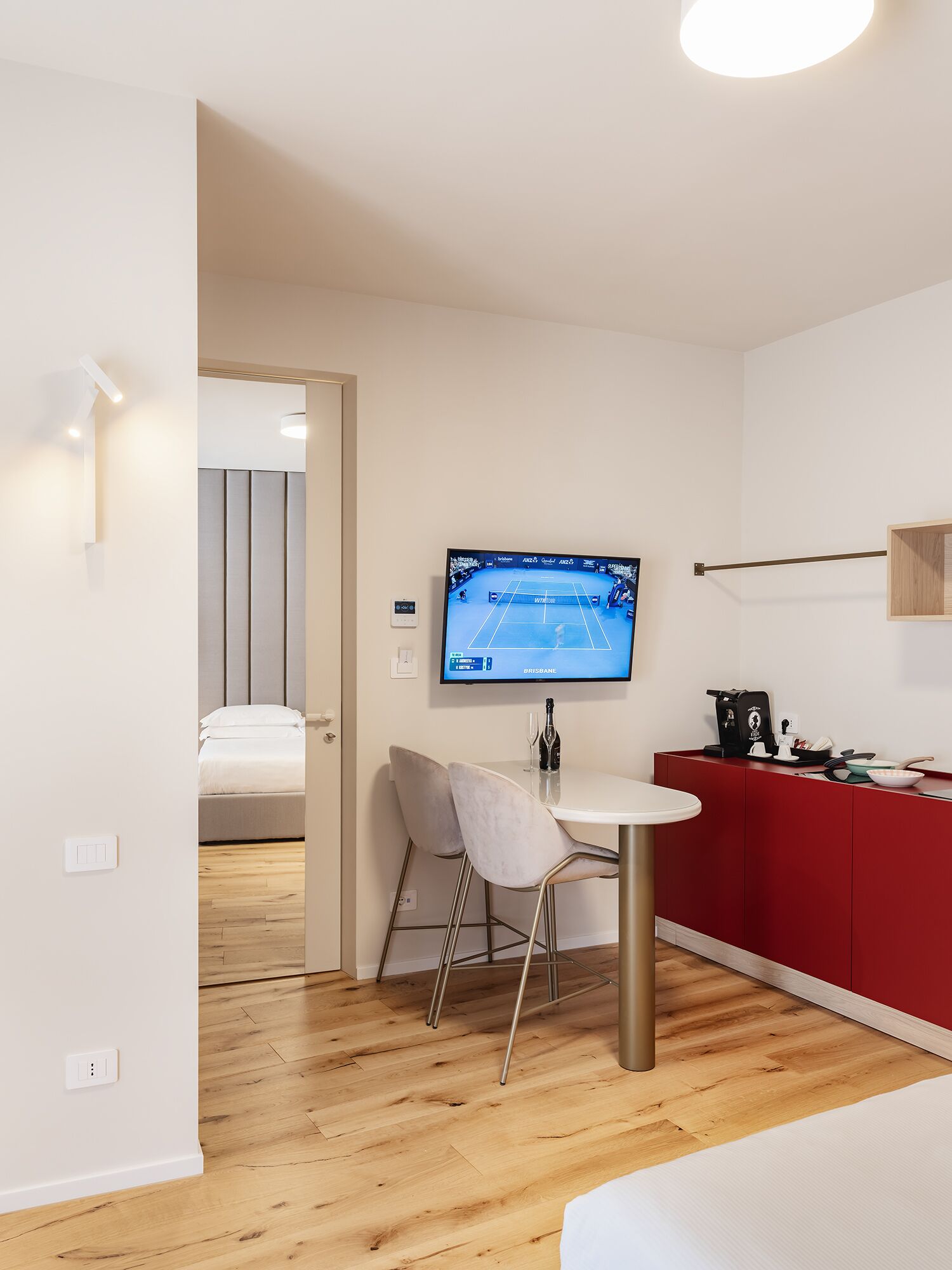 A modern room with a TV displaying a tennis match, a small dining area, and a red kitchenette.