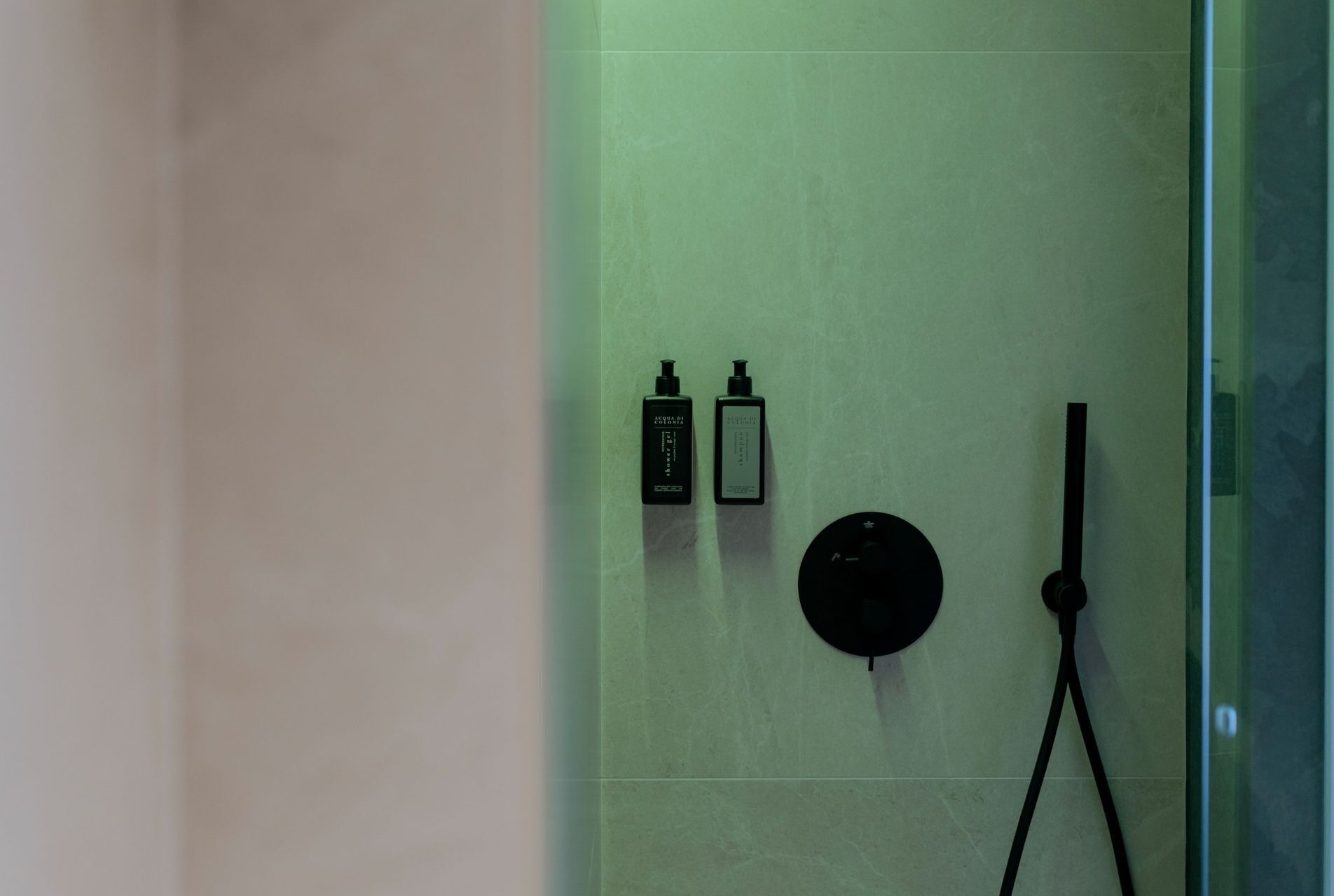 A bathroom with a green-tiled wall, two black bottles of Acqua di Colonia, a black circular object, and a black shower hose.