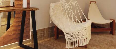 Una habitación con piso de madera, una silla colgante de macramé, una mesa de madera con un espejo y un techo con listones de madera.