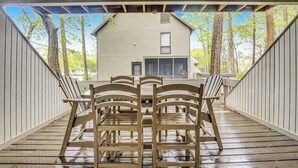 A covered outdoor deck with wooden chairs and tables, a view of a building in the background, and a wooden railing. - Sea Colony Tennis 1st-floor condo with balcony & tennis court (Bethany Beach)