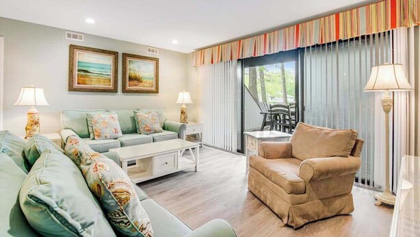A living room with a sofa, armchair, and coffee table, a dining area with chairs, and a view through large windows. - Sea Colony Tennis 1st-floor condo with balcony & tennis court (Bethany Beach)