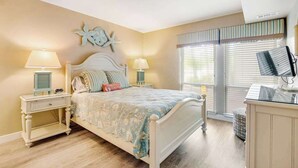 A bedroom with a bed, two bedside tables with lamps, a starfish decoration, a television, and a window with blinds. - Sea Colony Tennis 1st-floor condo with balcony & tennis court (Bethany Beach)