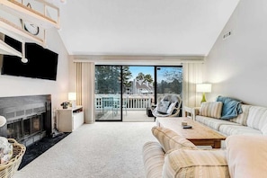 A spacious living room with a fireplace, a sofa, a coffee table, and a view through a glass door to an outdoor balcony. - Sea Colony 2nd Fl Condo Lake View, Pools, & Tennis Court (Bethany Beach)
