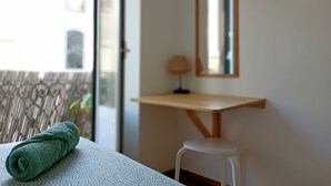 Une pièce comprenant un lit, un petit tabouret blanc, une table de chevet en bois, un miroir et une fenêtre donnant sur des immeubles.