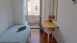 Une petite pièce comprenant un lit, un bureau, une chaise, un miroir et une vue sur l’extérieur grâce à une porte ouverte.