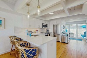 Una cocina moderna con armarios blancos, una isla central, taburetes de mimbre, electrodomésticos de acero inoxidable y un amplio ventanal.