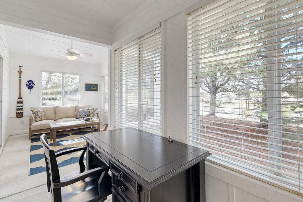 A room with a dining table, chairs, a sofa, and large windows with blinds.