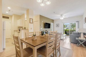 A modern kitchen with white cabinets, a dining table with chairs, a TV mounted on the wall, and a balcony with outdoor seating.