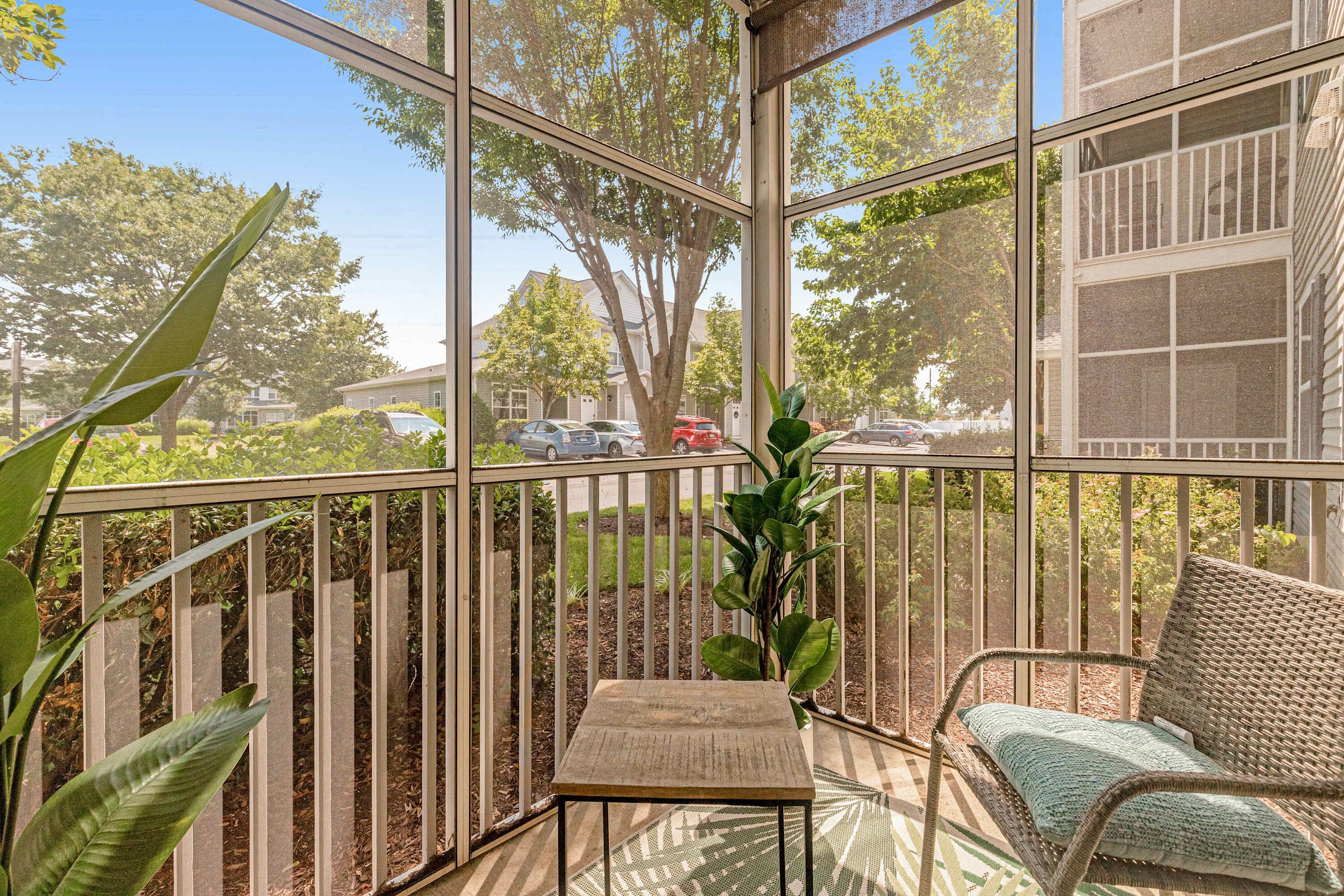 En solbeskinnet balkon med en fletstol og et lille bord, med udsigt over en boligvej og grøntområde.