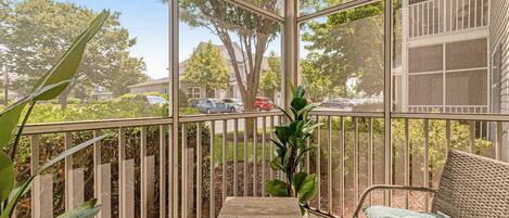 En solbeskinnet balkon med en fletstol og et lille bord, med udsigt over en boligvej og grøntområde.