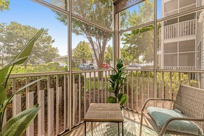 A sunlit balcony with a wicker chair and a small table, overlooking a residential street and greenery.