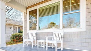Ein überdachter Balkon mit weißen Stühlen und einem kleinen Tisch, Holzverkleidung und ein Fenster mit Jalousien.