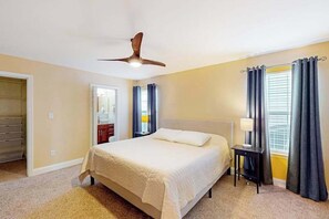 Ein Schlafzimmer mit einem Bett, einem Deckenventilator, einem Nachttisch mit Lampe und einem Fenster mit Vorhängen.