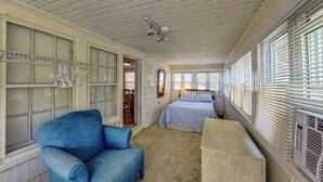 Een kamer met een bed, een blauwe stoel, een houten bank, een plafondventilator en grote ramen met jaloezieën.
