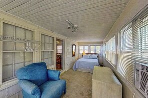 Une pièce comprenant un lit, une chaise bleue, un banc en bois, un ventilateur de plafond et de grandes fenêtres équipées de stores.