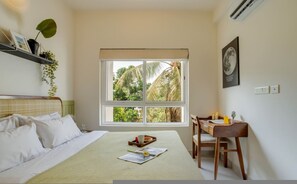 A bedroom with a bed, a desk, a window with a view of palm trees, a shelf with plants, and a framed picture on the wall. - Field View 2bhk Retreat 1km to Candolim Beach (Candolim)