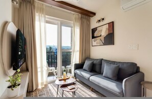 A living room with a grey sofa, a flat-screen TV, a potted plant, a small table with a bottle and glasses, and a balcony with a view. - Field View 2bhk Retreat 1km to Candolim Beach (Candolim)