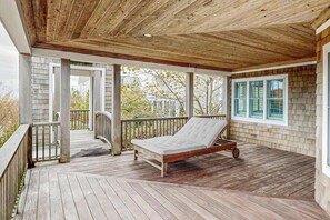 A covered porch with a wooden deck, a metal-framed bed, and a view of a house through a window. - The Chancellery pet friendly house with private patio, balcony & grill (Dewey Beach)