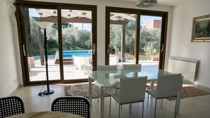 A dining area with a glass table and chairs, a view of a pool and garden through sliding glass doors, and a radiator on the wall. - Villa le Muse - beautiful detached villa with swimming pool, (Marsala)