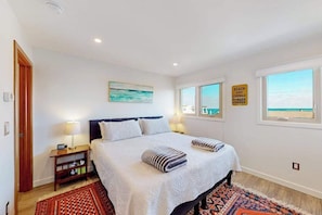 A bedroom with a bed, bedside tables, a rug, a painting, and a window with a view. - Lovely, ocean-block contemporary condo with pool, fast WiFi, balcony & W/D (Rehoboth Beach)