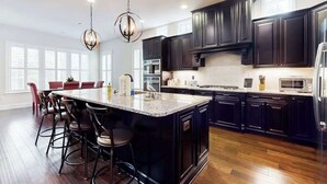 A modern kitchen with dark cabinetry, a large island with bar stools, and a chandelier. - Upgraded home with pools, sports courts, bikes, chef's kitchen, firepit, & gym (Rehoboth Beach)