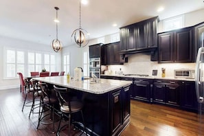 A modern kitchen with dark cabinetry, a large island with bar stools, and a chandelier. - Upgraded home with pools, sports courts, bikes, chef's kitchen, firepit, & gym (Rehoboth Beach)