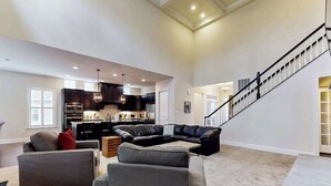 A spacious living room with a sectional sofa, a dining area, and a kitchen in the background. - Upgraded home with pools, sports courts, bikes, chef's kitchen, firepit, & gym (Rehoboth Beach)