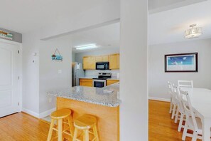 A kitchen with wooden cabinets, a granite countertop, and a dining area with white chairs. - Home near the ocean with a pool, outdoor space, WiFi, central AC, & washer/dryer (Rehoboth Beach)