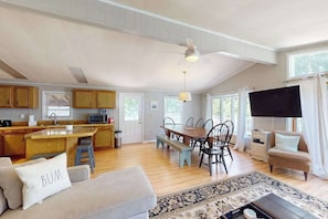 A spacious living area with a sofa, a dining table, and chairs, a kitchen with wooden cabinets, a ceiling fan, and a television. - Coastal Escape with Incredible Sunsets – Boat, Fish & Play at Angola By The Bay! (Lewes)