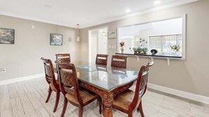 Comedor con mesa de vidrio rodeada de sillas de madera, un espejo grande y una ventana con plantas.