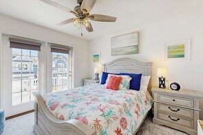 A bedroom with a bed, a ceiling fan, a nightstand, a lamp, and a window with a view of buildings. - Arbor-Lyn house with shared pool, fire pit, deck & gas grill (Rehoboth Beach)