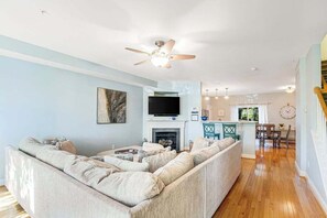 A living room with a sectional sofa, a flat-screen TV, a fireplace, and a dining area with a kitchen in the background. - Edgewater Park townhome with pool, patio, grill, balcony, AC & beach gear (Rehoboth Beach)