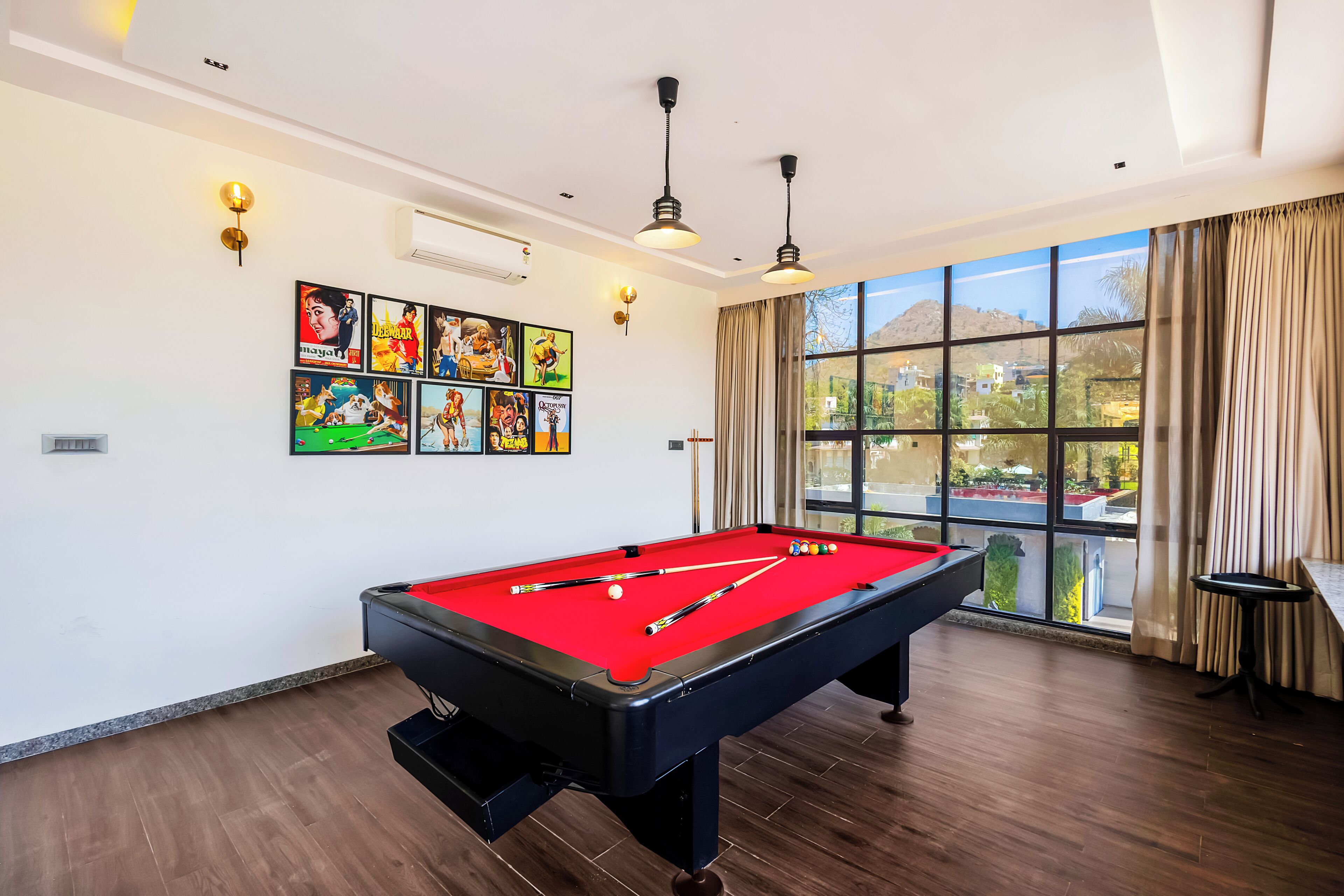 Une table de billard avec des billes, une queue de billard et une table de billard recouverte d’un feutre rouge. Il y a des lampes fixées au mur et une grande fenêtre avec des rideaux.