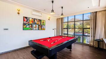 Une table de billard avec des billes, une queue de billard et une table de billard recouverte d’un feutre rouge. Il y a des lampes fixées au mur et une grande fenêtre avec des rideaux.