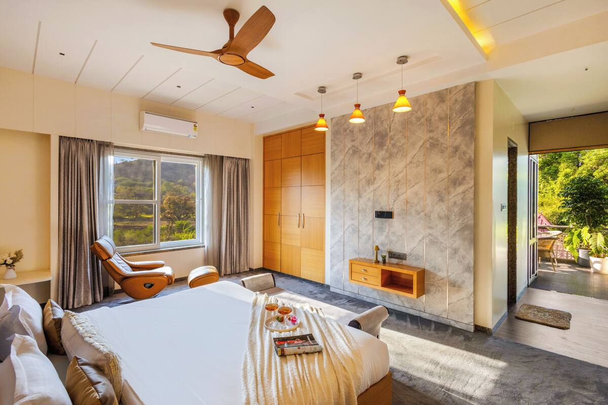 A spacious bedroom with a large bed, a ceiling fan, a wooden wardrobe, a marble wall, and a view of greenery outside.