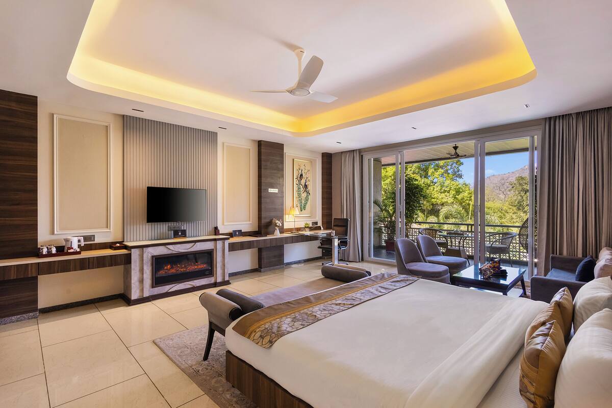 A modern hotel room with a large bed, a TV, a fireplace, a balcony with seating, and a ceiling fan.