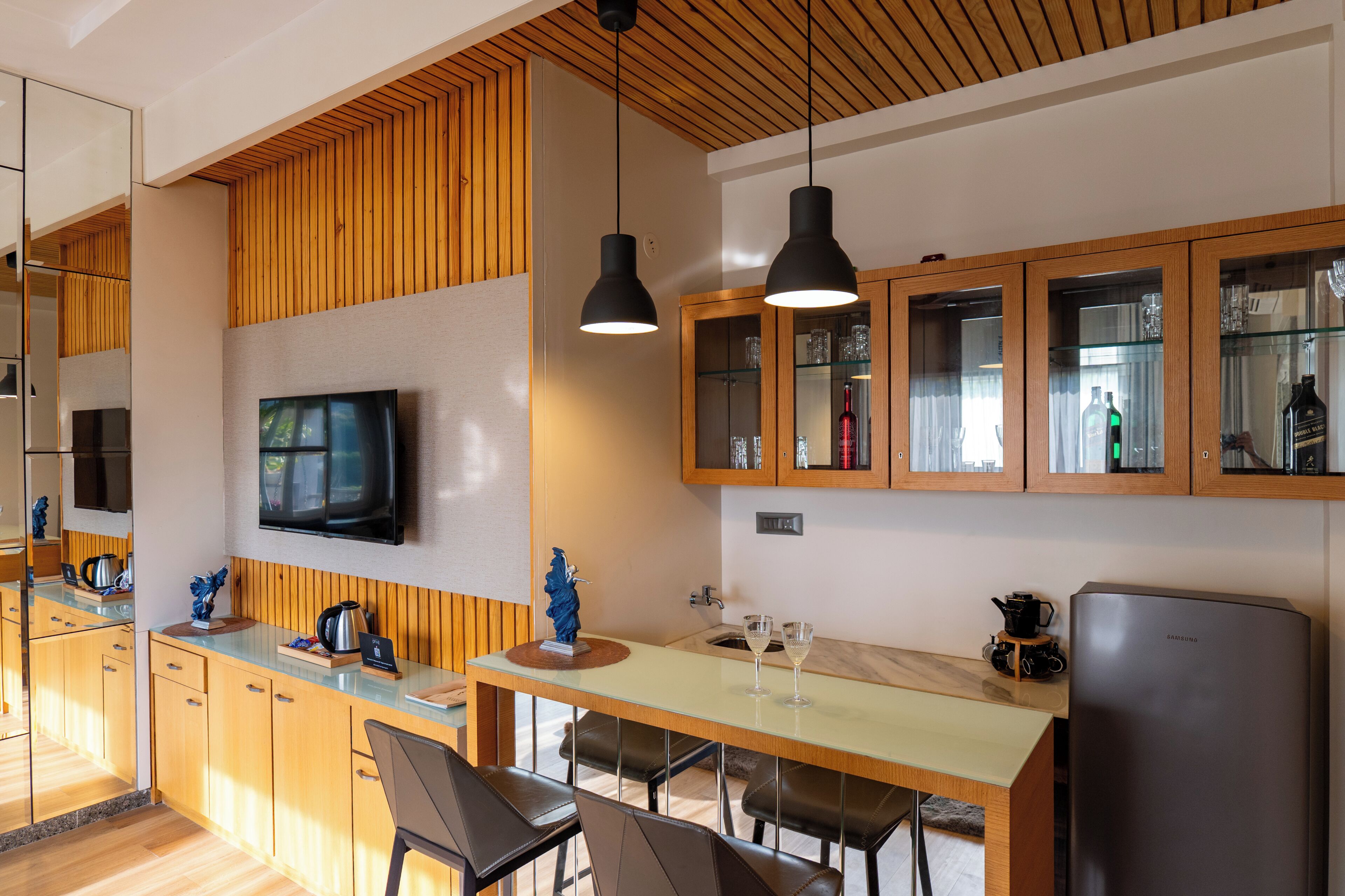 A modern kitchen with wooden cabinets, a flat-screen TV, pendant lights, and a refrigerator.
