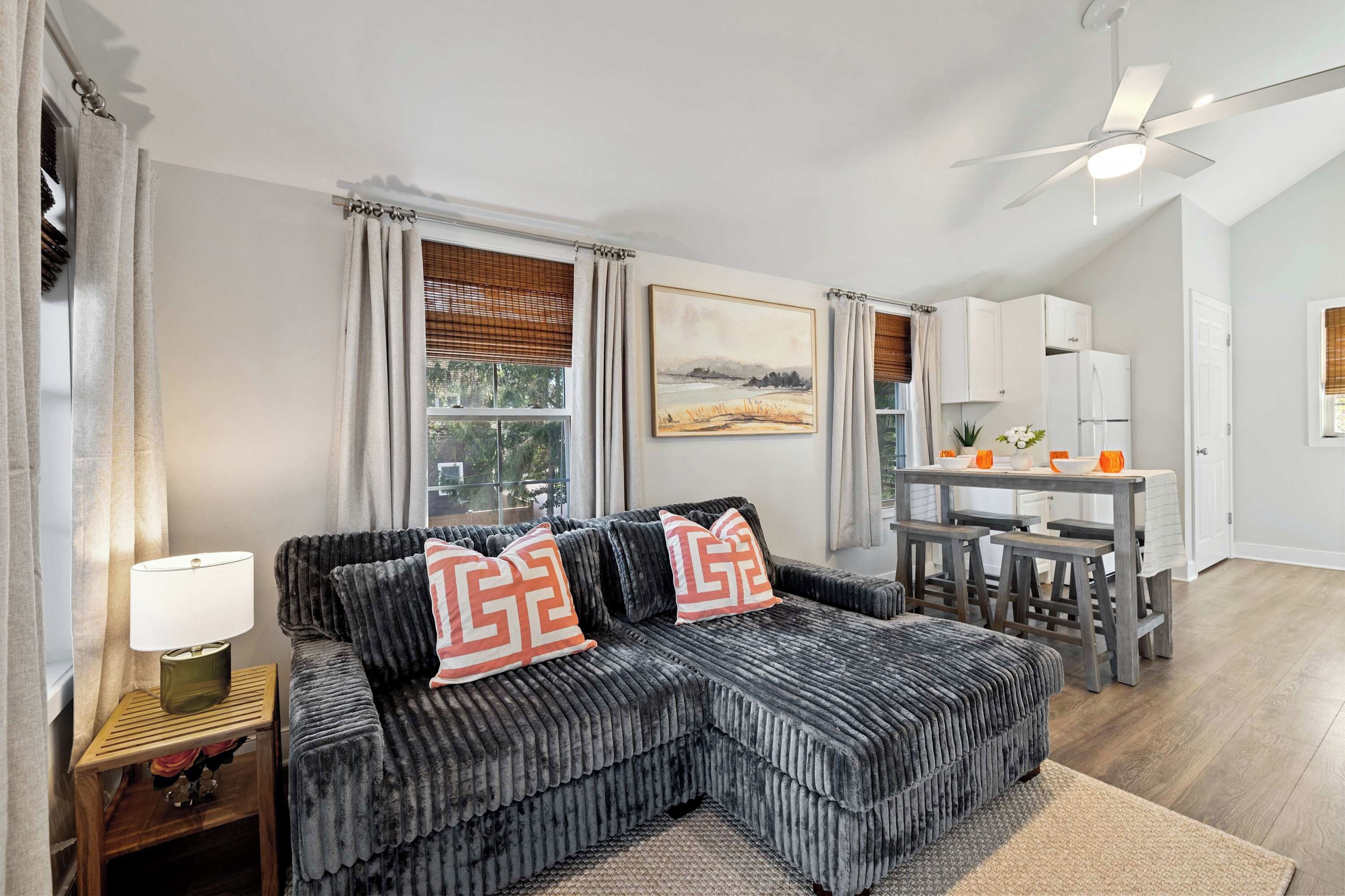 A modern living room with a sectional sofa, a dining area with bar stools, and a kitchen with white cabinets.