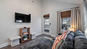 A living room with a sofa, a television mounted on the wall, a small wooden side table, and a door leading to a balcony.
