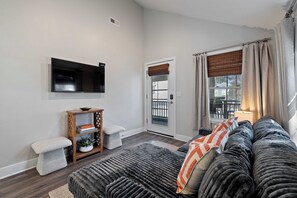 A living room with a sofa, a television mounted on the wall, a small wooden side table, and a door leading to a balcony.
