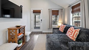 A living room with a sofa, a television, a wooden side table with books, and a door leading outside. - Dog-friendly Rehoboth Beach cottage steps from the beach & boardwalk (Rehoboth Beach)