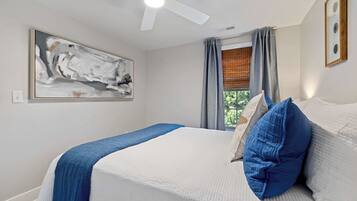 Une chambre à coucher avec un lit, un ventilateur de plafond, une fenêtre avec des rideaux et un tableau accroché au mur.
