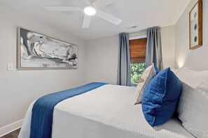 A bedroom with a bed, a ceiling fan, a window with curtains, and a painting on the wall.