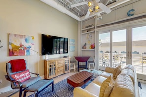 A modern living room with a sofa, a coffee table, a TV, a wooden cabinet, and a balcony with outdoor furniture. - Rehoboth Beach dog friendly townhome w/deck, patio, grill close to marina (Rehoboth Beach)