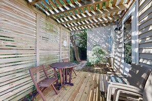 Una terraza de madera con pérgola, una mesa con sillas y un sillón de mimbre.