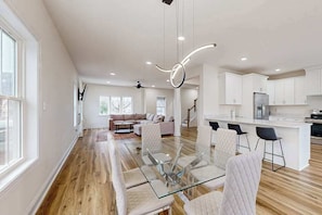 A modern kitchen with white cabinets, a central island, and a dining area with glass-top table and cushioned chairs. - Spacious townhouse w/patio, balcony, WIFI, W/D (Rehoboth Beach)