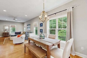 A spacious living room with a wooden dining table, white sofas, and a large window with curtains. - Town of Rehoboth Beach cottage with covered porch, deck, grill, AC, & W/D (Rehoboth Beach)