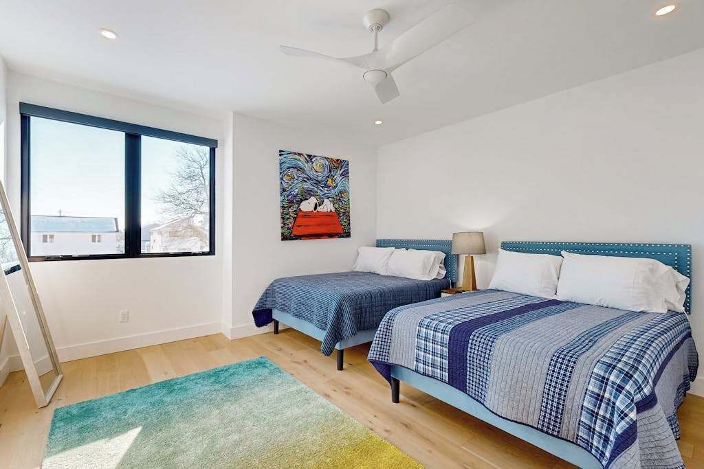 A bedroom with two beds, a ceiling fan, a window with a view, and a painting on the wall.