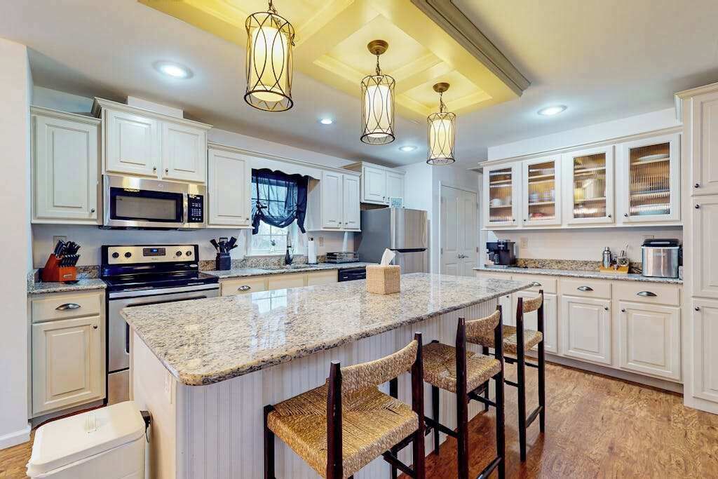 Una cocina con armarios blancos, encimera de granito, una isla central con taburetes de bar e iluminación colgante.